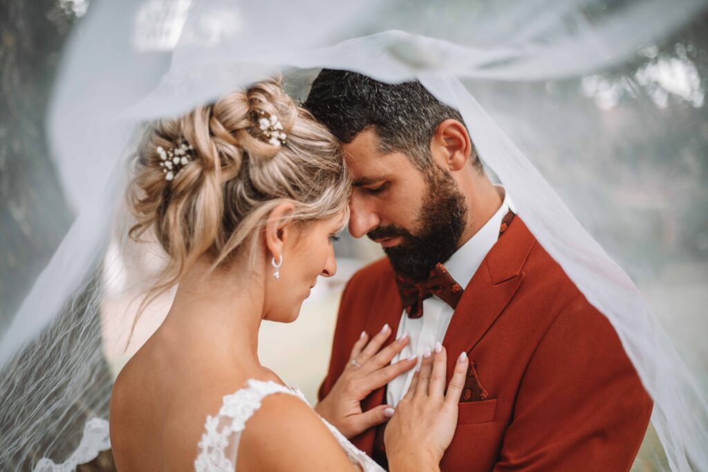 Sous un voile blanc, les mariés se tiennent côte à côte, front contre front, main dans la main. La mariée porte une robe blanche ornée de dentelle, tandis que le marié porte un costume couleur rouille avec un nœud papillon. photographe Normandie