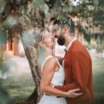 Une mariée en robe blanche et un marié en costume couleur rouille partagent un moment joyeux et intime sous un arbre feuillu le jour de leur mariage, souriant et s'embrassant à l'extérieur. photographe Normandie