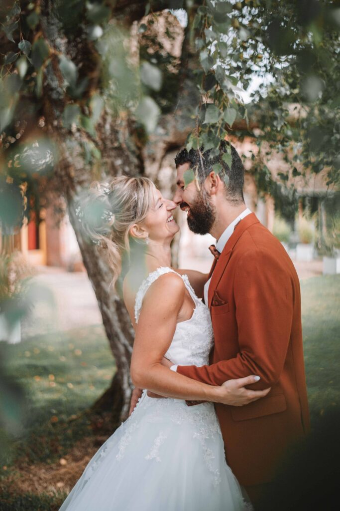 Une mariée en robe blanche et un marié en costume couleur rouille partagent un moment joyeux et intime sous un arbre feuillu le jour de leur mariage, souriant et s'embrassant à l'extérieur. photographe Normandie