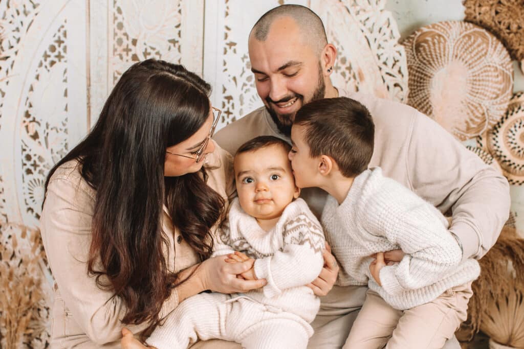 Une famille de quatre personnes est assise, souriante. Une femme tient un bébé aux grands yeux et à l'air sérieux, tandis qu'un homme est assis à côté d'elles. Un enfant plus âgé se penche pour embrasser le bébé sur la joue. Tous portent des vêtements clairs. photographe Normandie