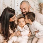 Une famille de quatre personnes est assise, souriante. Une femme tient un bébé aux grands yeux et à l'air sérieux, tandis qu'un homme est assis à côté d'elles. Un enfant plus âgé se penche pour embrasser le bébé sur la joue. Tous portent des vêtements clairs. photographe Normandie