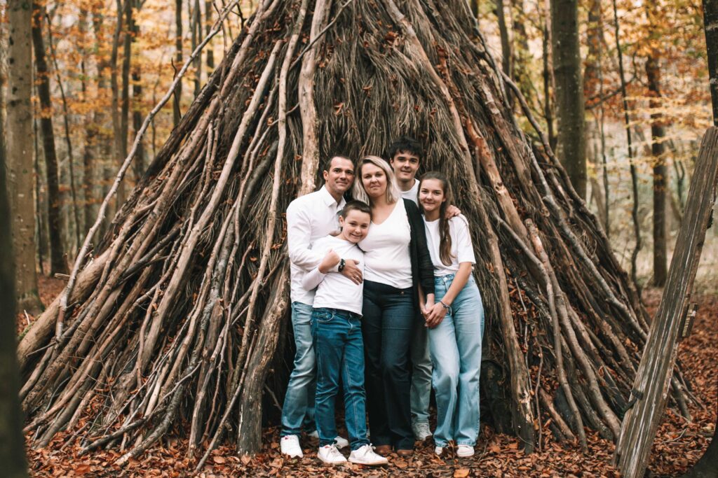 Une famille de cinq personnes, tous vêtus de hauts blancs et de jeans, se tient souriante devant une grande structure de tipi faite de branches dans une forêt aux feuilles d'automne. photographe Normandie