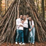 Une famille de cinq personnes, tous vêtus de hauts blancs et de jeans, se tient souriante devant une grande structure de tipi faite de branches dans une forêt aux feuilles d'automne. photographe Normandie