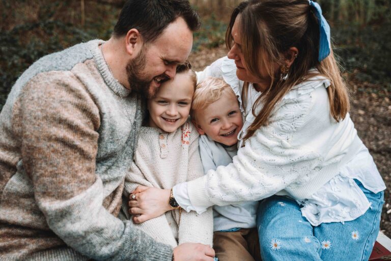 Une famille de quatre personnes est assise à l'extérieur, les bras croisés, souriant et riant. Les parents serrent leurs deux jeunes enfants, une fille et un garçon, dans leurs bras, créant un moment chaleureux et joyeux. photographe Normandie