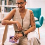 Femme assise sur une table dans une bibliothèque habillé en beige tien un livre dans ses mains et pose regard vers l'objectif Séance photo professionnel