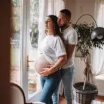 couple avec une femme enceinte s'enlace dans une belle piece normande l'homme avec la main sur le ventre de la femme les 2 habillé en haut blanc et jean. Photographe grossesse Normandie