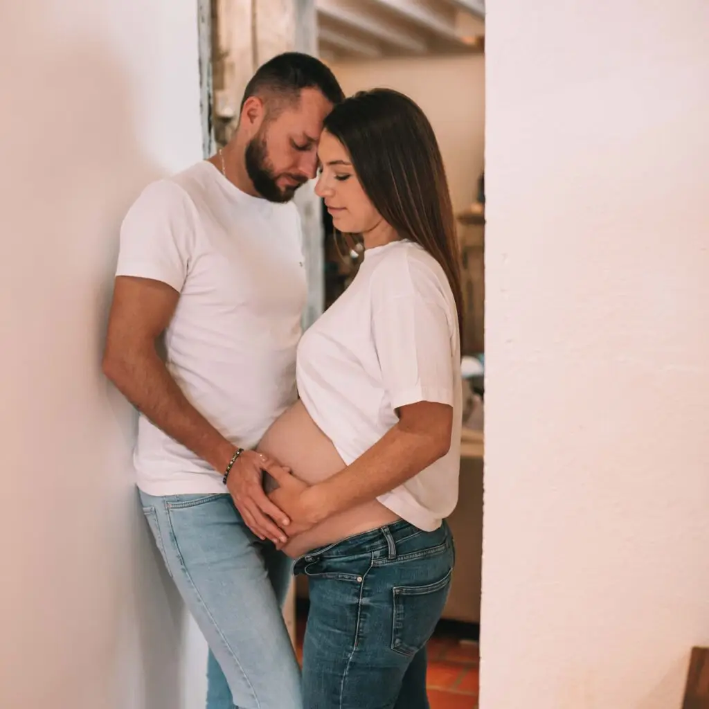 couple avec une femme enceinte s'enlace dans une belle piece normande l'homme avec la main sur le ventre de la femme les 2 habillé en haut blanc et jean. Photographe grossesse Normandie