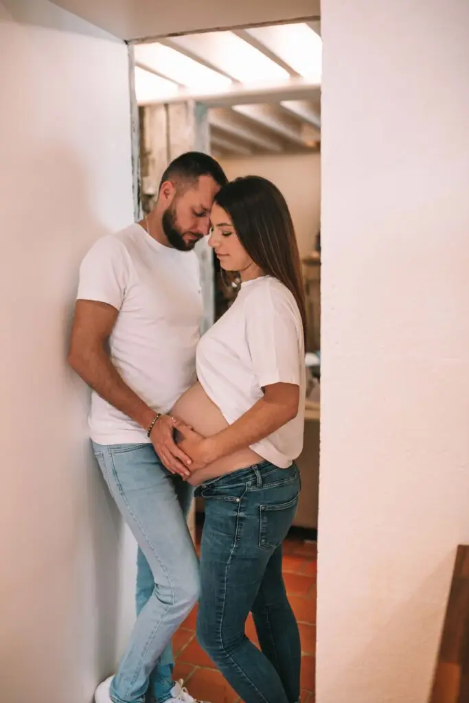 couple avec une femme enceinte s'enlace dans une belle piece normande l'homme avec la main sur le ventre de la femme les 2 habillé en haut blanc et jean. Photographe grossesse Normandie