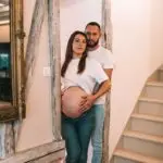 couple avec une femme enceinte s'enlace dans une belle piece normande avec poutre en bois l'homme avec la main sur le ventre de la femme les 2 habillé en haut blanc et jean. Photographe grossesse Normandie