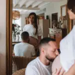 couple avec une femme enceinte s'enlace dans une belle piece normande dans le reflet d'un miroir l'homme embrasse le ventre de la femme les 2 habillé en haut blanc et jean. Photographe grossesse Normandie