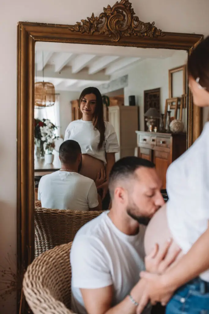 couple avec une femme enceinte s'enlace dans une belle piece normande dans le reflet d'un miroir l'homme embrasse le ventre de la femme les 2 habillé en haut blanc et jean. Photographe grossesse Normandie