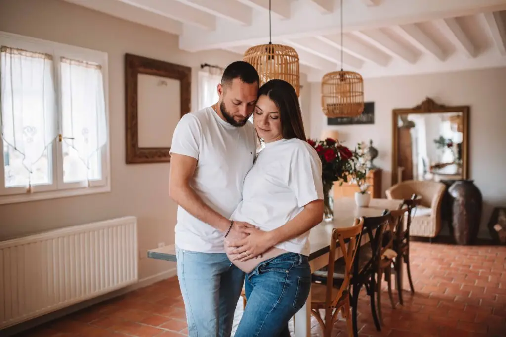 couple avec une femme enceinte s'enlace dans une belle piece normande l'homme avec la main sur le ventre de la femme les 2 habillé en haut blanc et jean. Photographe grossesse Normandie