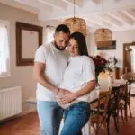 couple avec une femme enceinte s'enlace dans une belle piece normande l'homme avec la main sur le ventre de la femme les 2 habillé en haut blanc et jean. Photographe grossesse Normandie