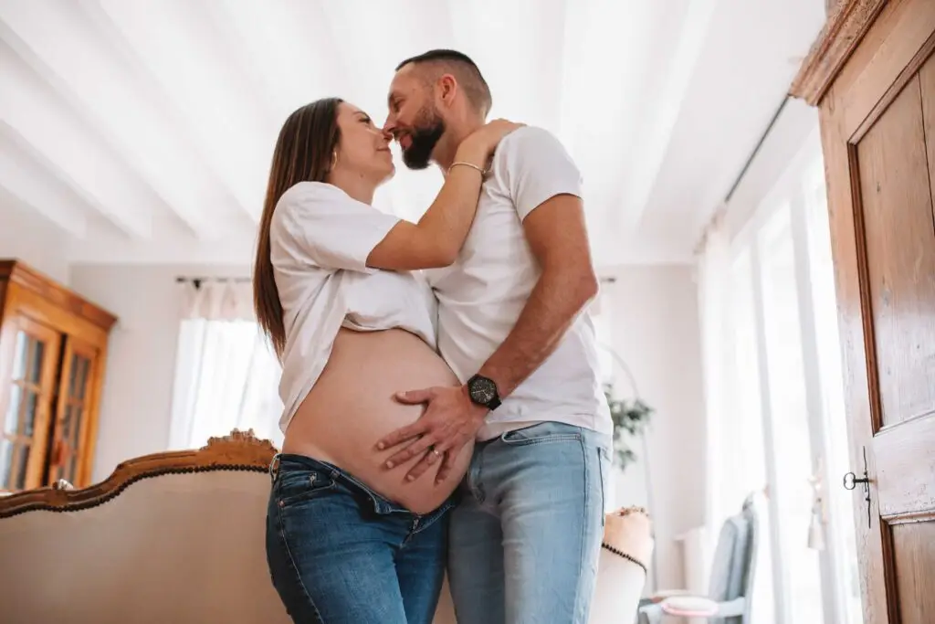 couple avec une femme enceinte s'enlace dans une belle piece normande l'homme avec la main sur le ventre de la femme les 2 habillé en haut blanc et jean. Photographe grossesse Normandie