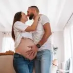 couple avec une femme enceinte s'enlace dans une belle piece normande l'homme avec la main sur le ventre de la femme les 2 habillé en haut blanc et jean. Photographe grossesse Normandie
