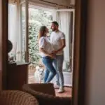 couple avec une femme enceinte s'enlace devant une fenetre, on voit leur reflet dans le miroir et un petit chien blanc accompagne l'homme avec la main sur le ventre de la femme les 2 habillé en haut blanc et jean. Photographe grossesse Normandie