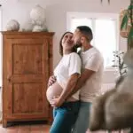 couple avec une femme enceinte s'enlace dans une belle piece normande l'homme avec la main sur le ventre de la femme les 2 habillé en haut blanc et jean. Photographe grossesse Normandie