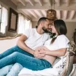 couple avec une femme enceinte s'enlace dans une belle piece normande assis sur un canapé d'epoque l'homme avec la main sur le ventre de la femme les 2 habillé en haut blanc et jean. Photographe grossesse Normandie