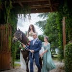 Une mariée en robe blanche monte un cheval noir mené par un homme en costume bleu, avec une femme en robe verte marchant à côté d'eux, le tout sous un portail rustique couvert de vigne à l'extérieur. photographe Normandie