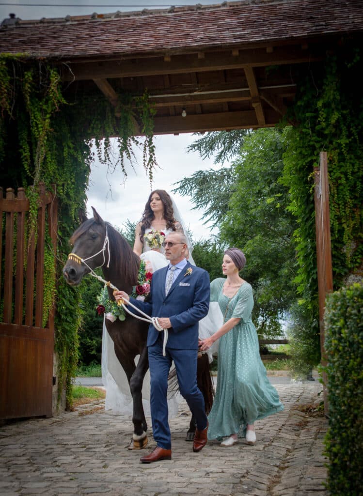 Une mariée en robe blanche monte un cheval noir mené par un homme en costume bleu, avec une femme en robe verte marchant à côté d'eux, le tout sous un portail rustique couvert de vigne à l'extérieur. photographe Normandie