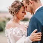 Un couple de mariés s'embrassent en plein air, front contre front, souriant doucement. La mariée porte une robe blanche en dentelle à épaules dénudées et détails floraux ; le marié porte un costume bleu. La scène est tendre et romantique. photographe Normandie