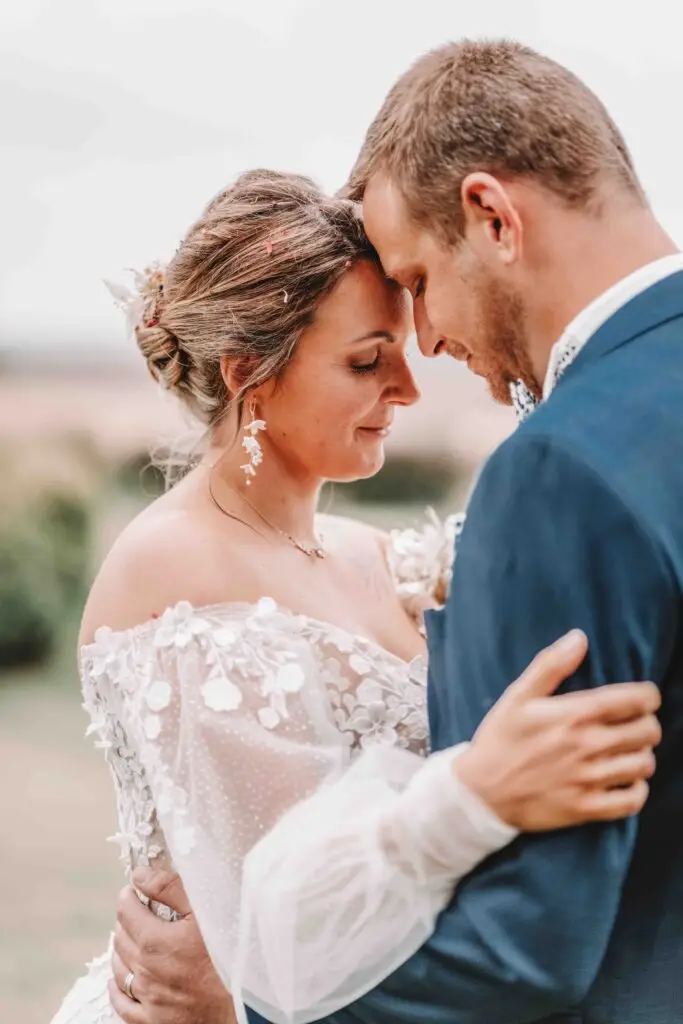 Un couple de mariés s'embrassent en plein air, front contre front, souriant doucement. La mariée porte une robe blanche en dentelle à épaules dénudées et détails floraux ; le marié porte un costume bleu. La scène est tendre et romantique. photographe Normandie