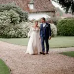 Une mariée en robe blanche et un marié en costume bleu marchent main dans la main sur un chemin de gravier, se souriant, avec de la verdure et un bâtiment rustique en arrière-plan. photographe Normandie