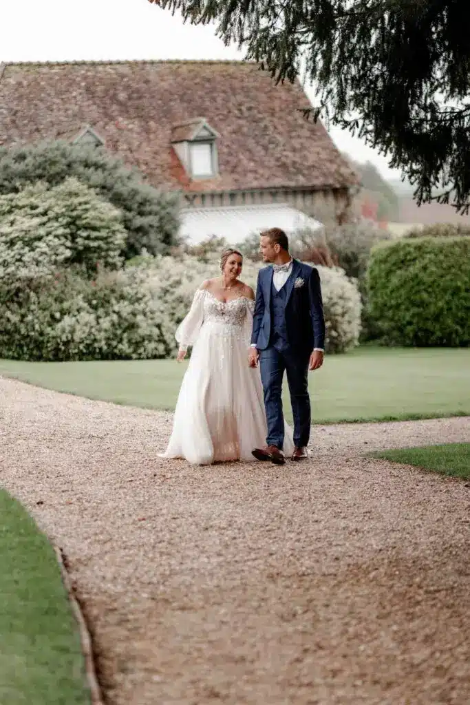 Une mariée en robe blanche et un marié en costume bleu marchent main dans la main sur un chemin de gravier, se souriant, avec de la verdure et un bâtiment rustique en arrière-plan. photographe Normandie