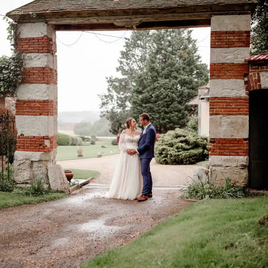 Une mariée et un marié se tiennent face à face sous une arche rustique en brique et en pierre, entourés de verdure et d'un chemin de gravier, partageant un moment intime le jour de leur mariage. photographe Normandie