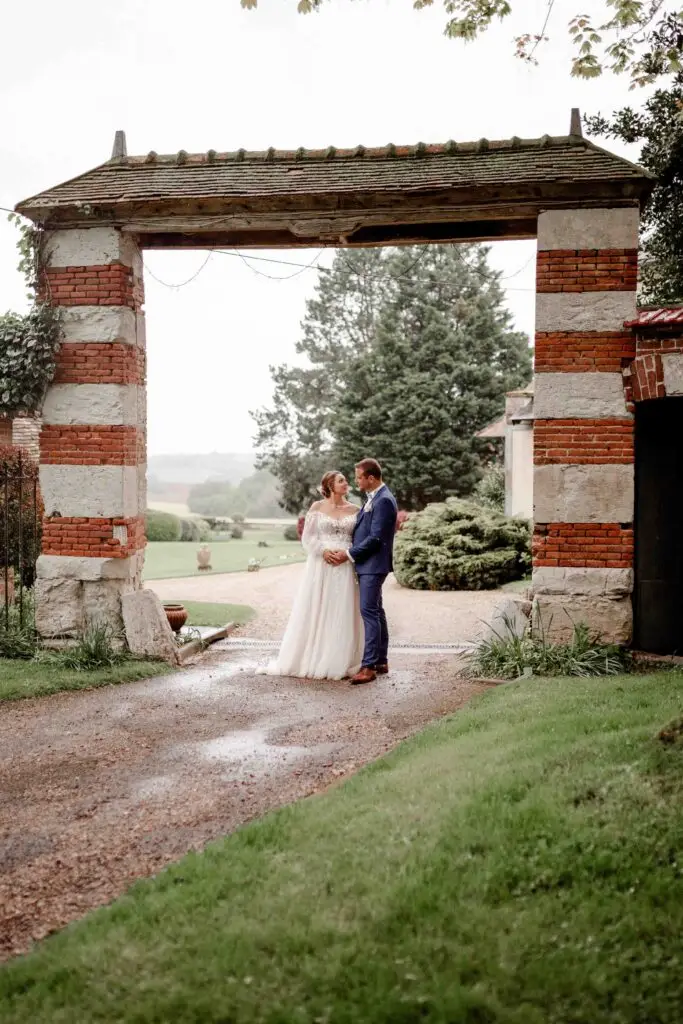 Une mariée et un marié se tiennent face à face sous une arche rustique en brique et en pierre, entourés de verdure et d'un chemin de gravier, partageant un moment intime le jour de leur mariage. photographe Normandie