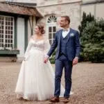 Une mariée vêtue d'une robe blanche en dentelle à manches longues et un marié vêtu d'un costume bleu se tiennent la main à l'extérieur d'un bâtiment de style vintage, se regardant et souriant. photographe Normandie