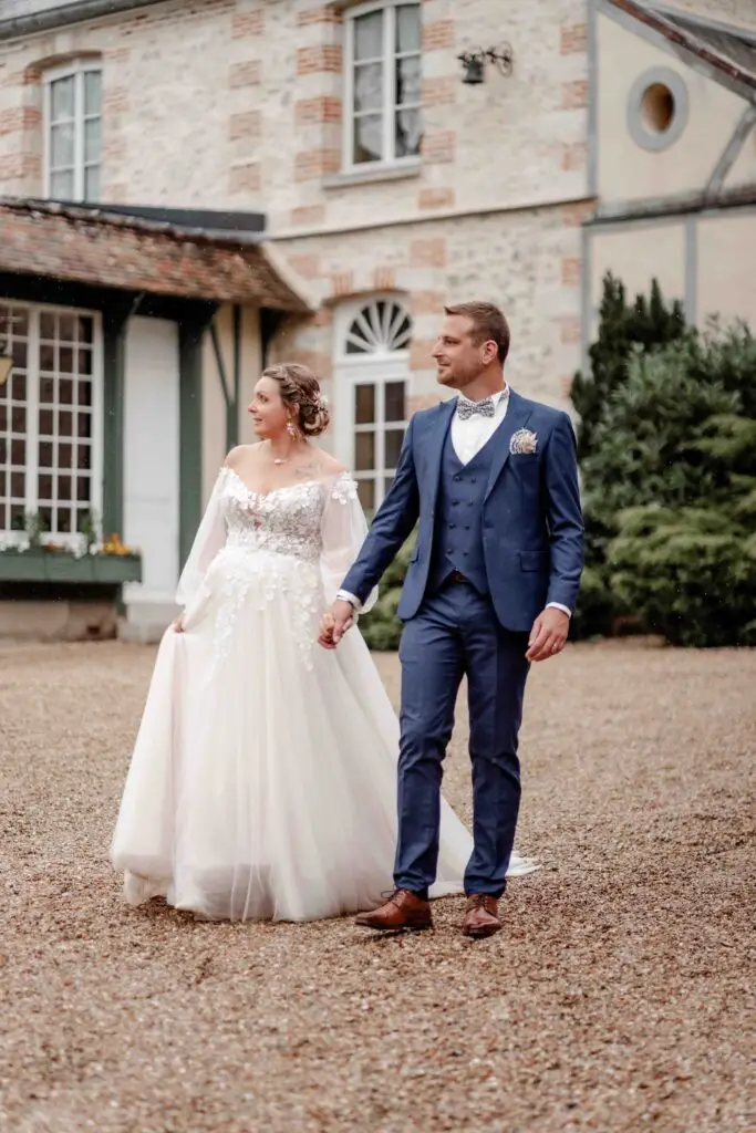 Une mariée vêtue d'une robe blanche en dentelle à manches longues et un marié vêtu d'un costume bleu se tiennent la main à l'extérieur d'un bâtiment de style vintage, se regardant et souriant. photographe Normandie
