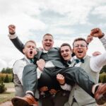 Quatre hommes en costume applaudissent joyeusement en plein air, tenant un homme souriant au centre. Ils semblent célébrer, peut-être un mariage, avec de la verdure et un ciel nuageux en arrière-plan. photographe Normandie