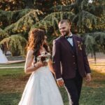 Une mariée en robe de mariée blanche et un marié en costume rayé se tiennent la main et se sourient tout en marchant dehors sur une pelouse herbeuse, avec de grands arbres en arrière-plan. photographe Normandie
