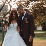 Une mariée en robe blanche, bouquet à la main, marche dehors avec un marié en costume rayé sombre. Ils sourient et se penchent l'un vers l'autre, entourés d'arbres et d'un soleil radieux. photographe Normandie