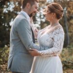 Un couple de mariés se tient debout dehors, face à face, souriant et se tenant tendrement dans leurs bras. La mariée porte une robe en dentelle blanche et le marié un costume gris clair. La lumière du soleil filtre à travers les arbres en arrière-plan. photographe Normandie