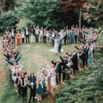 Un grand groupe de personnes se tient dehors, sur l'herbe, formant un cœur, le visage tourné vers l'intérieur, les mains levées en signe de célébration. Des arbres et des buissons verdoyants entourent le rassemblement. photographe Normandie