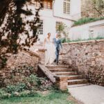 Une mariée en robe blanche et un homme en costume bleu descendent des marches en pierre à l'extérieur d'une grande maison élégante avec de hautes fenêtres, entourée de verdure et d'un mur de pierre par une journée ensoleillée. photographe Normandie
