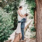 Un marié en costume bleu soulève sa mariée en robe blanche tandis qu'ils se sourient, entourés d'arbres verts luxuriants et de feuillages par une journée ensoleillée. photographe Normandie