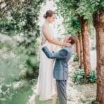 Une mariée en robe blanche se tient sur un rebord de pierre, regardant vers le bas et souriant à un marié en costume bleu qui lui tient la taille, entouré de verdure et d'arbres dans un jardin extérieur. photographe Normandie