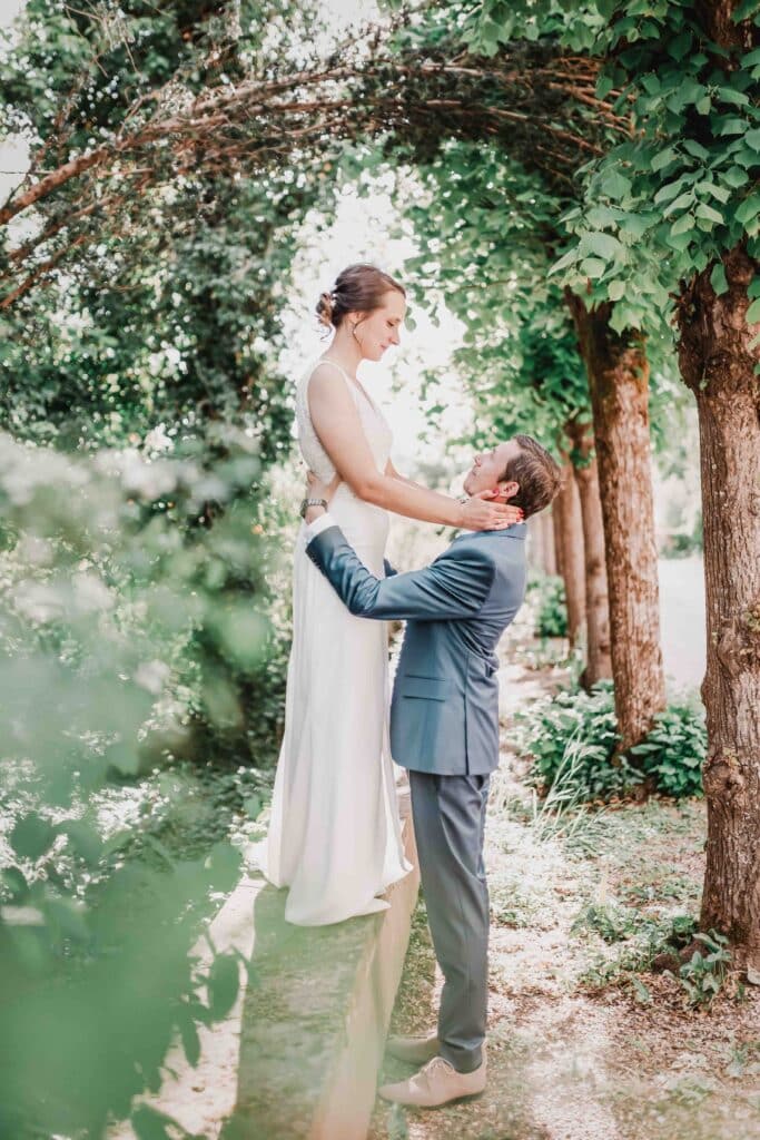 Une mariée en robe blanche se tient sur un rebord de pierre, regardant vers le bas et souriant à un marié en costume bleu qui lui tient la taille, entouré de verdure et d'arbres dans un jardin extérieur. photographe Normandie